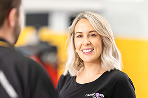 A Capricorn Risk Account Manager smiling while engaging in conversation with a Member in a workshop setting, with a blurred background featuring automotive equipment and yellow accents.